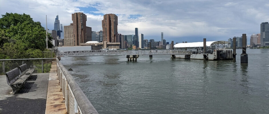 Waterfront walkway and ferry terminal with NYC skyline backdrop.