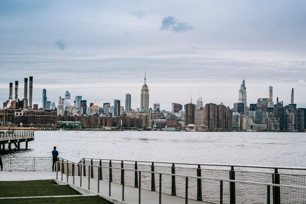 Man looking at Manhattan skyline from a pier across water.