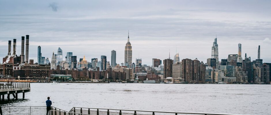 Man looking at Manhattan skyline from a pier across water.