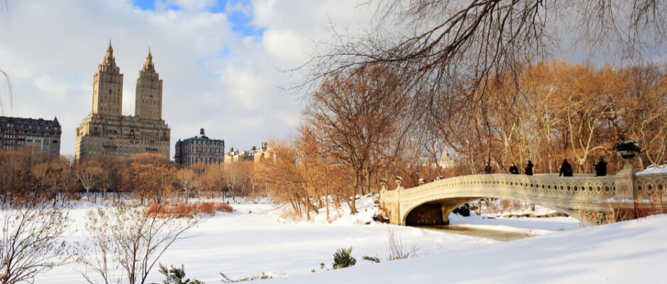 Snow-covered Bow Bridge in Central Park with city skyline background.