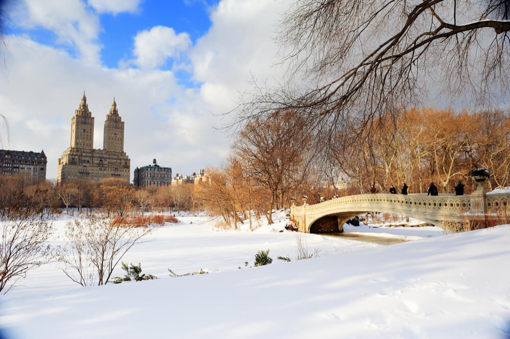 Snow-covered Bow Bridge in Central Park with city skyline background.